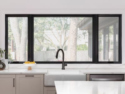 Kitchen remodel with a black faucet on a white farmhouse sink, black framed windows, cabinets, and white marble countertops.