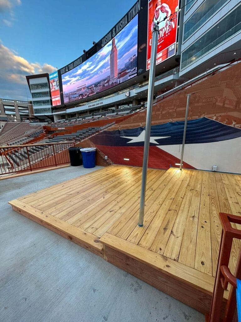 A close-up view of the custom wooden deck at DKR-Texas Memorial Stadium, showcasing the natural wood finish and open space with stadium seating and the scoreboard in the background.