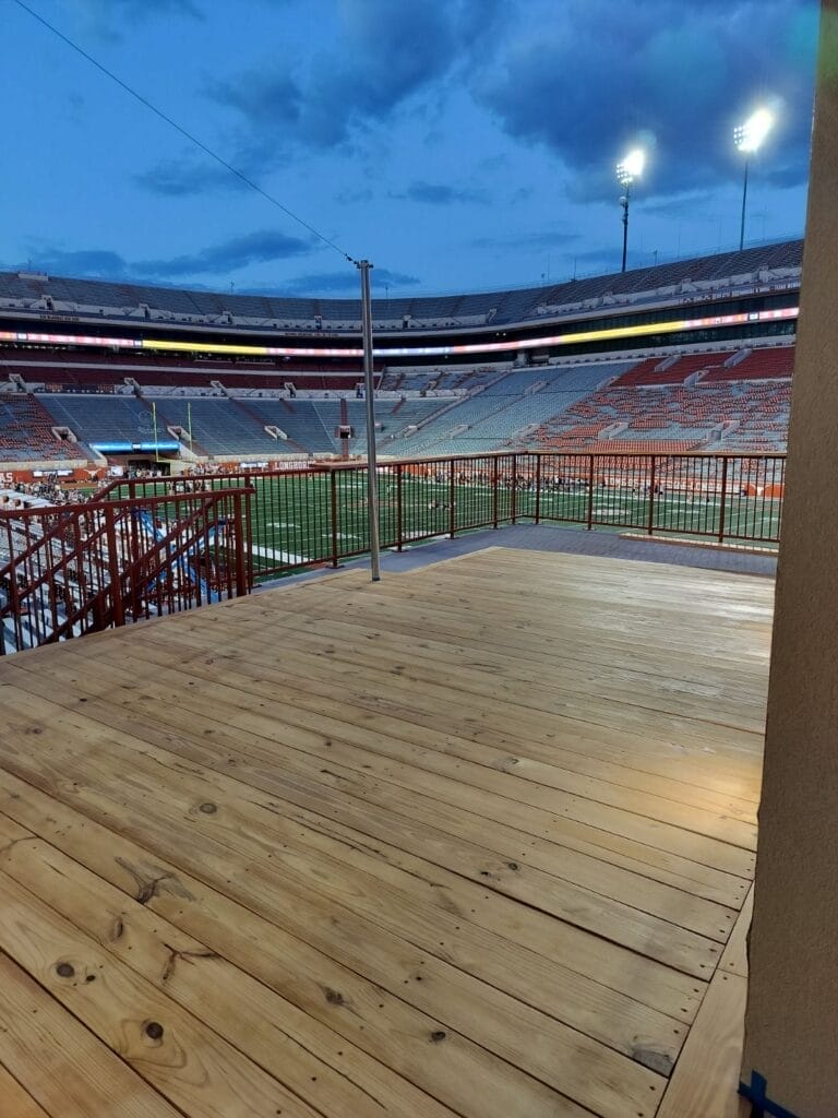 A wide view of the wooden deck at dusk inside Darrell K Royal-Texas Memorial Stadium, with the stadium lights illuminating the field.