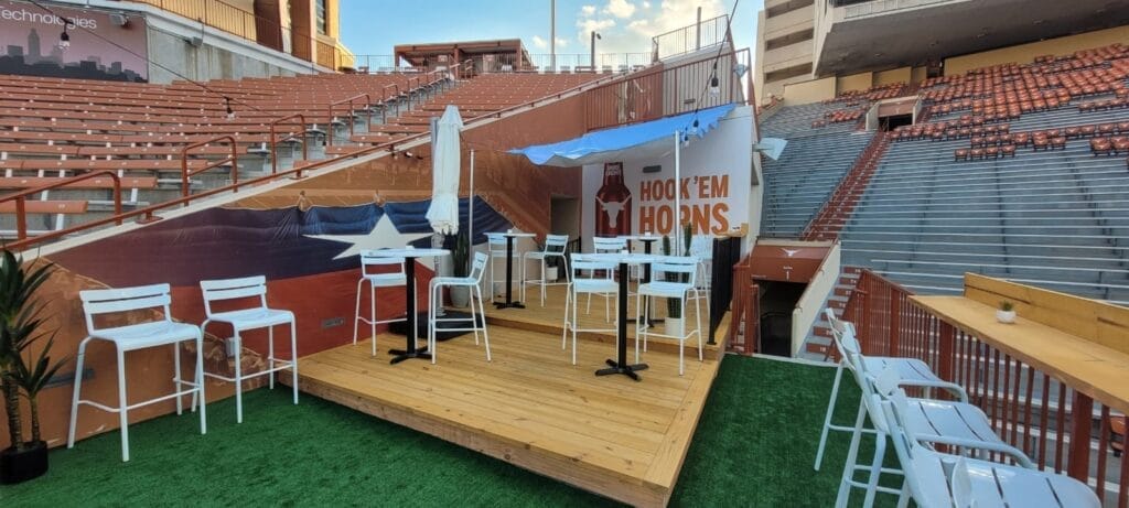 A newly constructed wooden deck inside Darrell K Royal-Texas Memorial Stadium featuring high-top tables and chairs, with a view of the stadium's stands in the background.