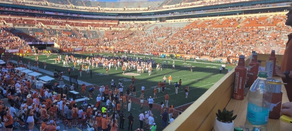 A game-day view from the custom-built deck overlooking the crowded field at Darrell K Royal-Texas Memorial Stadium, filled with fans dressed in burnt orange for a Texas Longhorns football game.