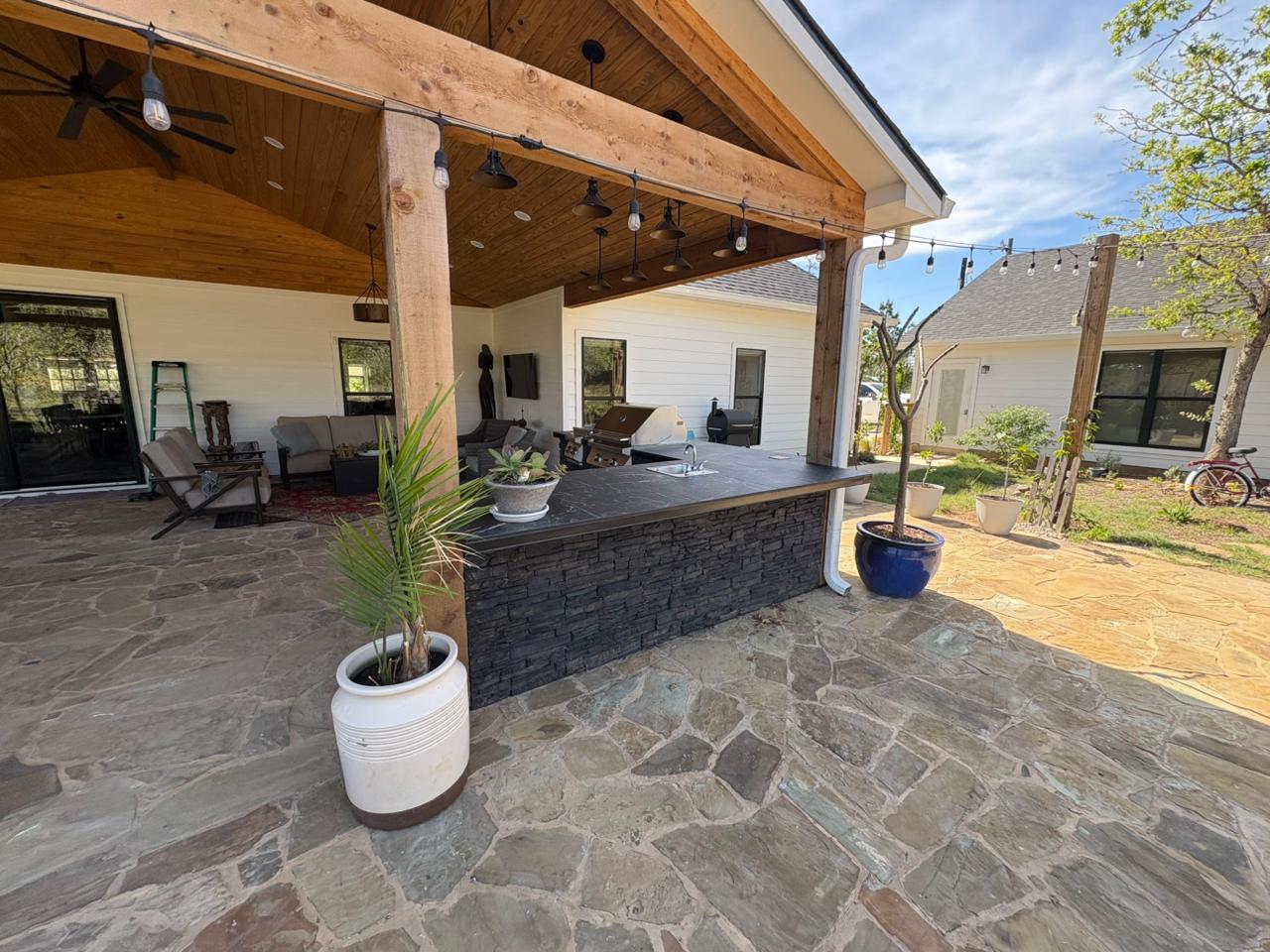 Wide view of covered patio outdoor kitchen with L-shaped stacked stone island, bar seating, string lights, and flagstone patio in Bastrop, TX
