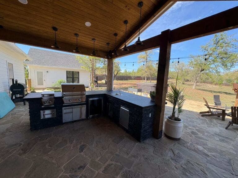 Custom outdoor kitchen with stacked stone island, built-in grill, storage drawers, pendant barn lights, and tongue-and-groove wood ceiling under a covered patio in Bastrop