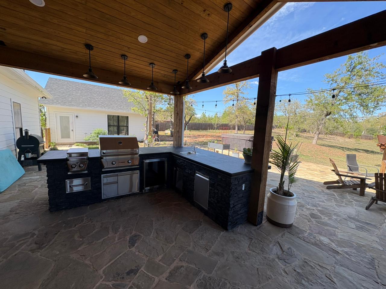 Custom outdoor kitchen with stacked stone island, built-in grill, storage drawers, pendant barn lights, and tongue-and-groove wood ceiling under a covered patio in Bastrop
