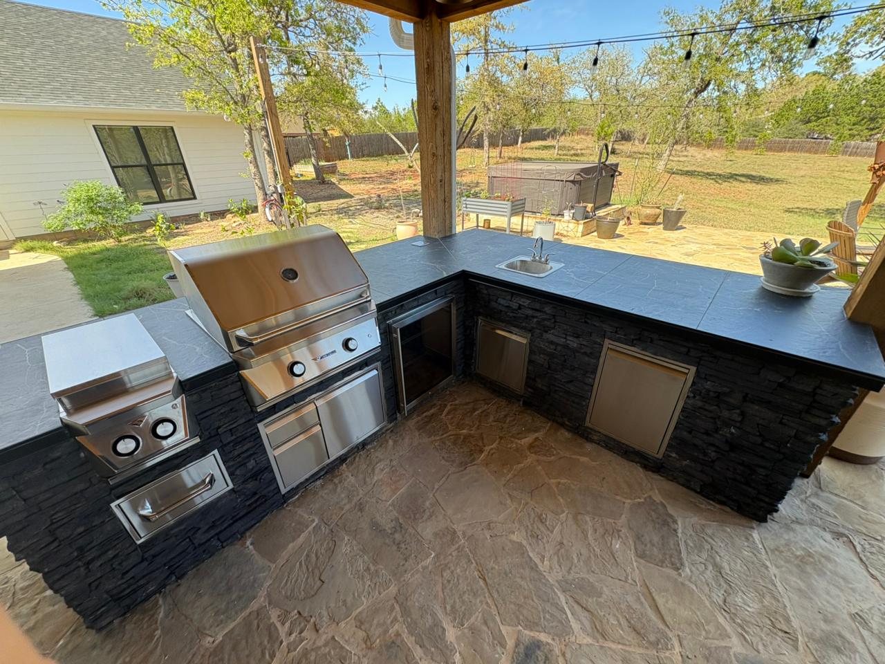 Close-up of outdoor kitchen working side showing built-in stainless steel grill, side burner, sink, access doors, and refrigerator with dark stacked stone veneer in Bastrop