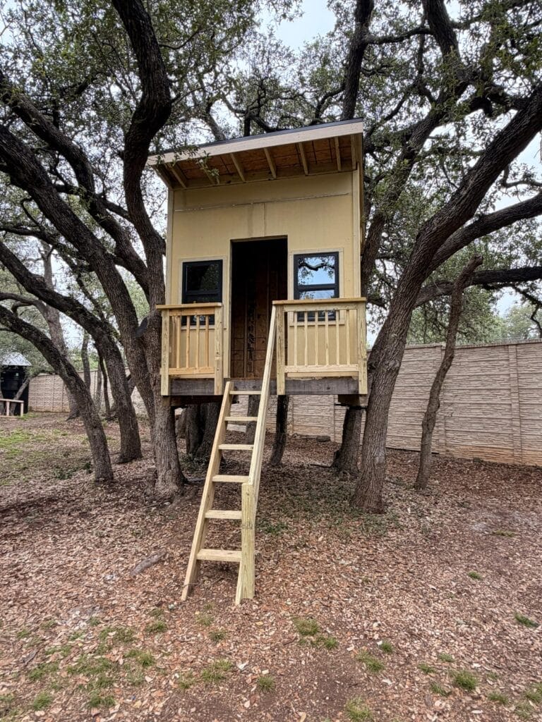 Front view of custom treehouse showing black-framed windows, balustered porch, and lean-to roof