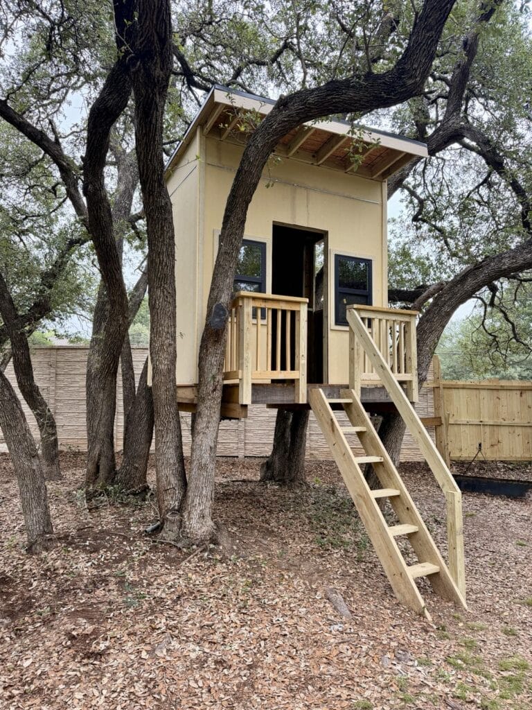 Front view of custom treehouse showing black-framed windows, balustered porch, and lean-to roof